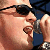A close-up of a person with light skin wearing dark sunglasses and a blue sleeveless top, holding a silver phone to their ear.