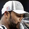 Static close-up of a person wearing a white and gray baseball cap with visible logos, holding a silver trophy or cup near their face against a dark background.