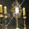 Static icon depicting a golden monstrance with radiating light beams and three yellow candles in ornate holders against a dark background.