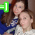 Two people in a close pose with one person behind the other, wearing blue and white clothing, with a green, purple, and pink emoticon symbol in the top-left corner.
