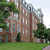 A pixelated static image shows a multi-story brick building with tall windows, surrounded by green trees and grass under a light sky.