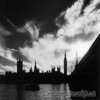 Black-and-white silhouette of a city skyline with spires and towers reflected in water under a cloudy sky.
