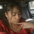A person in a red shirt makes a peace sign with their right hand while seated inside a vehicle, with a blurred background visible through the window.