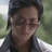 Pixelated static image of a person with dark hair wearing glasses and a dark jacket over a light shirt, set against an outdoor background.