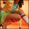 A person in a crouched pose wearing a yellow jacket, light blue shirt, green shorts, and white knee-high socks with red text and black-red stripes.