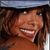 A static pixelated image shows a person with long brown hair smiling broadly, wearing a brown top against a dark background with light blue and white elements at the top.