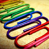 Colorful paper clips in rainbow order on a textured beige surface, arranged in a diagonal line from top-left to bottom-right.