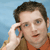 Pixelated image of a person holding a blue phone to their ear against a light blue background, wearing an orange shirt and brown jacket.