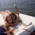 A person in a bikini lies on a white towel by water, with one arm raised and the other resting near their chest.