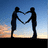 Two silhouetted figures stand against a gradient sky, holding hands to form a heart shape with their arms.