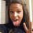 Pixelated close-up of a person with long brown hair wearing a dark top, making a hand gesture near their face.