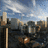 Cityscape with tall buildings under a blue sky with white clouds, featuring a bright light source in the lower center.
