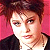 Pixelated portrait of a person with short brown hair against a pink background, wearing dark clothing and a light-colored shirt.