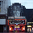 Pixelated image of a building facade with dark upper structure, red-framed lower section, and illuminated windows.
