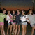 Group of people standing together in a line, wearing various colored shirts against a dark background.