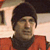 Pixelated image of a person with dark hair wearing a red top and dark collar against a light background with framed objects.