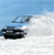 Pixelated image of a dark vehicle on a light surface under a blue sky with a blurred background element.