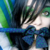 A close-up of a person with dark hair, green eyeshadow, and a blue polka-dot bow covering their mouth against a striped background.