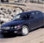 A dark blue sedan with visible headlights and wheels is positioned on a light-colored road, with a blurred background of buildings and sky.