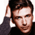 Static pixelated image of a person with dark hair wearing a dark long-sleeve shirt, hand raised to head in a gesture against a white background.