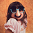 Pixelated static image of a person with dark hair and glasses wearing a light-colored top against an orange background.