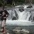 Pixelated static image showing a figure with dark hair and light clothing standing near a body of water with a bridge in the background.
