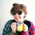A person with brown hair and sunglasses holds a yellow object in front of their face, wearing a multicolored checkered sweater against a light background.