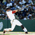 A baseball player in a white uniform with red accents swings a bat on a field with a green outfield wall and blurred spectators.