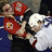 Two hockey players in red and white jerseys collide on ice with a bright red background displaying blue text.