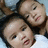 Two people with dark hair and light skin pose closely together against a blurred background, one person's head resting on the other's shoulder.