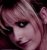 Pixelated close-up of a person's face with light skin tone, dark eyes, and pinkish hair against a dark background.