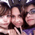 Three people with dark hair pose closely together in a static image, their faces partially visible against a blurred background.