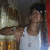 A person in a white tank top and necklace poses with a hand gesture against a blurred background with warm lighting.