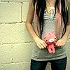 A person with long dark hair holds a small pink plush toy against a light-colored brick wall background.