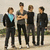 Four young men stand in a against an outdoor backdrop, with one wearing a light blue shirt under his dark jacket