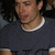 Pixelated close-up of a person wearing dark clothing with white trim, showing partial face and neck against a dark background.