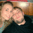 Two people in close physical proximity, one with light hair and the other with dark hair, both wearing dark clothing against a blurred background.