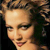 Pixelated close-up of a person's face with light skin tone, dark curly hair, and a neutral expression against a dark background.