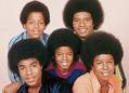 Static image of five young Black men with afro hairstyles posing together in a group portrait, wearing colorful casual clothing against a plain light background.