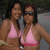 Two women in pink bikini tops pose together, one wearing sunglasses, against a blurred outdoor background.