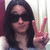 A person with long dark hair and sunglasses makes a peace sign, wearing a light-colored top against a striped background.