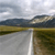 A static pixelated landscape image showing a road with a white line, grassy fields, distant mountains, and a cloudy sky.