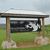 A static pixelated image shows a wooden shelter structure with a flat roof supported by four posts, set against a grassy field and light blue sky.