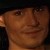 Pixelated close-up of a person's face with warm brown skin tone, visible eye, nose, and mouth, dark background, and partial shoulder.