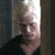 Pixelated static image of a person with light hair wearing dark clothing, positioned against a bright vertical light source on the right side.
