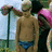 Pixelated static image shows a shirtless person in blue swimwear standing outdoors with a streetlamp and pink flowers visible in the background.