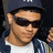 Pixelated static image of a person wearing dark sunglasses, a white shirt, and a dark jacket with blue accents.