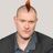 Pixelated static image of a person with a mohawk hairstyle wearing a dark jacket over a light shirt against a light background.