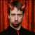 Static pixelated image of a person with dark hair and small red devil horns against a vertical striped red background, wearing a black shirt with a visible red tie or collar detail.