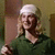 A person with long blonde hair and a white headband wears a dark green polo shirt, holding an object in their right hand against a striped background.